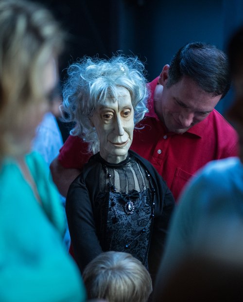Mężczyzna stoi obok lalki teatralnej. Lalka naturalnych rozmiarów, odwzorowana w sposób rzeczywisty, przedstawia starszą kobietę. Jej twarz jest wyrzeźbiona w drewnie.