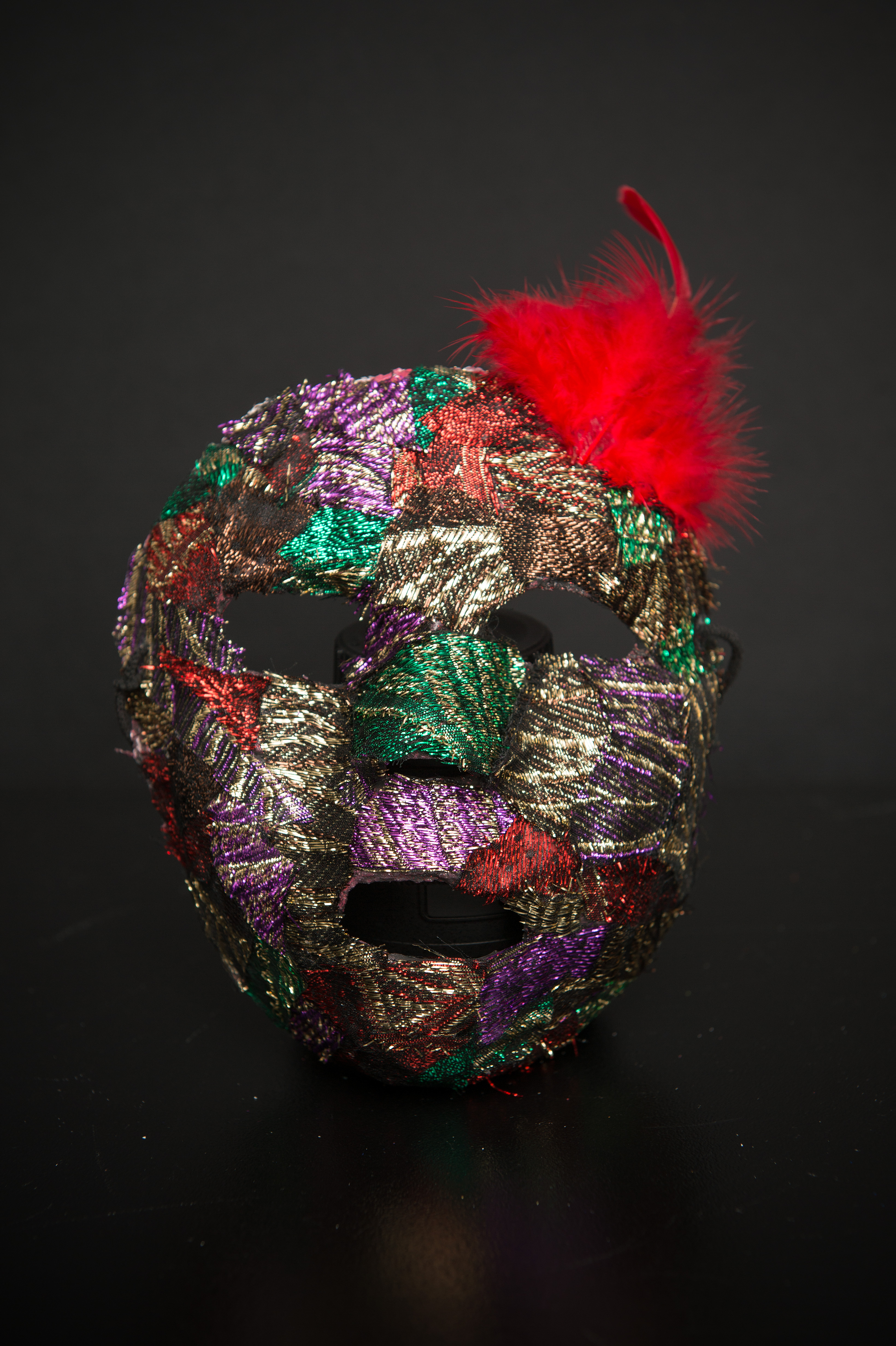Maska teatralna, bardzo kolorowa, z doczepionym u góry czerwonym piórem.