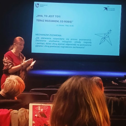 Prowadząca i uczestnicy siedzący na widowni, w tle ekran z projekcją