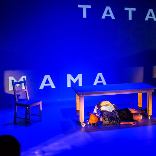 Spektakl Tata. Napisy na ścianie MAMA i TATA. Aktorzy leżą na podłodze pod drewnianym stołem.