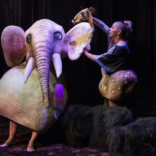 Spod kostiumu Słonia wystają gołe nogi aktora. Obok aktorka w kostiumie sarny wyciera serwetką ucho Słonia.