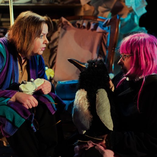Dwie aktorki siedzą naprzeciwko, zdjęcie w bliskim planie. Aktorka uchatka w szlafroku, mówi coś w ożywieniu. Aktorka pingwin zwrócona do niej, tuż przed jej twarzą trzyma puchatą lalkę pingwina.