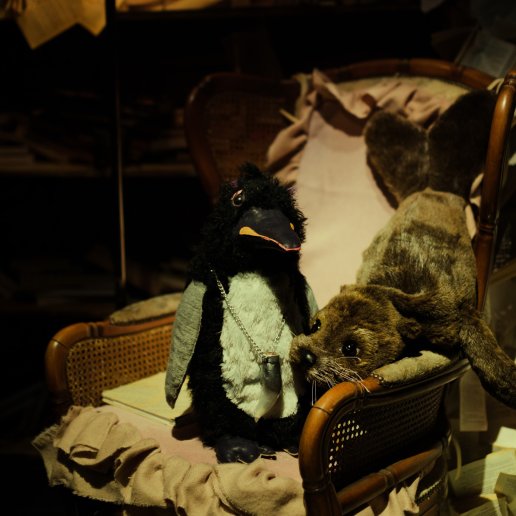 Na ratanowym krześle siedzą dwie lalki: pingwina i uchatki. Lalki są wykonane z puszystych materiałów, jak maskotki. Pingwin ma zawieszony na szyi naszyjnik z kłem. W tle świeci się lalka z materiałowym abażurem.