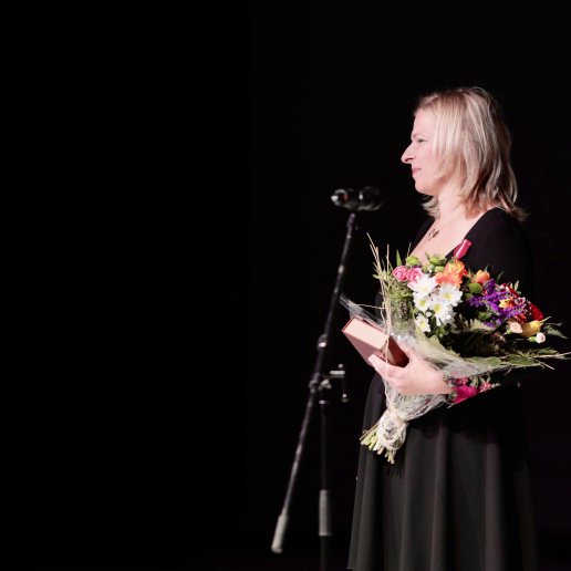 Agnieszka Zyskowska-Biskup stroi na scenie przy mikrofonie, z bukietem kwiatów. Ma półdługie blond włosy, jest uśmiechnięta i szczęśliwa. Ma sukienkę czarną w wyszywane, kolorowe kwiaty.
