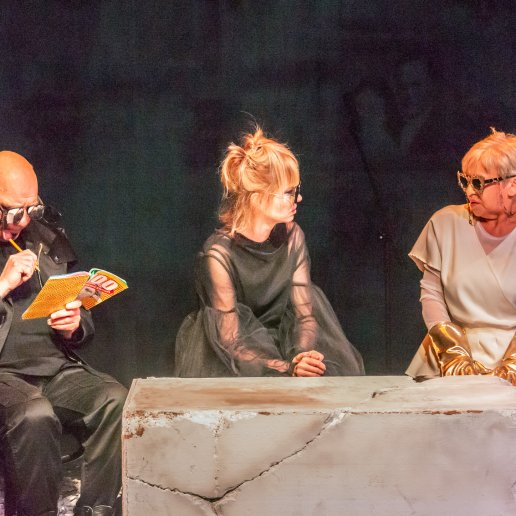 Spektakl Troja. Wojny nie będzie!. Trzech aktorów siedzi przy scenografii - ruinach miasta. Mają nietypowe okulary przeciwsłoneczne. Aktor rozwiązuje krzyżówkę.