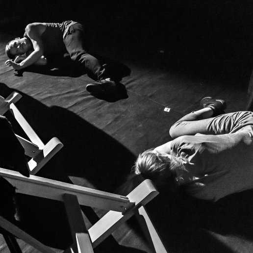 Skuleni aktorzy leżą naprzeciwko siebie na scenicznej podłodze. Widoczny fragment sztalug.