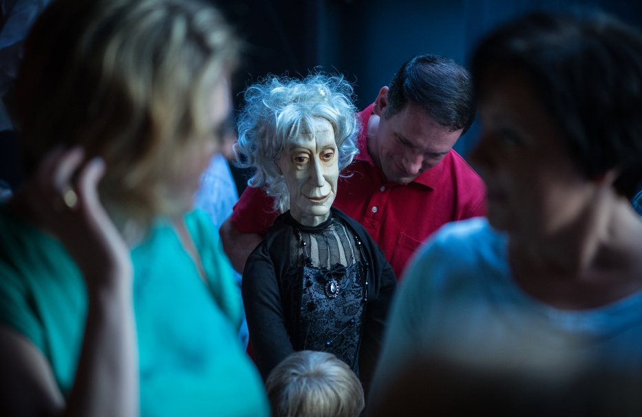 Mężczyzna stoi obok lalki teatralnej. Lalka naturalnych rozmiarów, odwzorowana w sposób rzeczywisty, przedstawia starszą kobietę. Jej twarz jest wyrzeźbiona w drewnie.