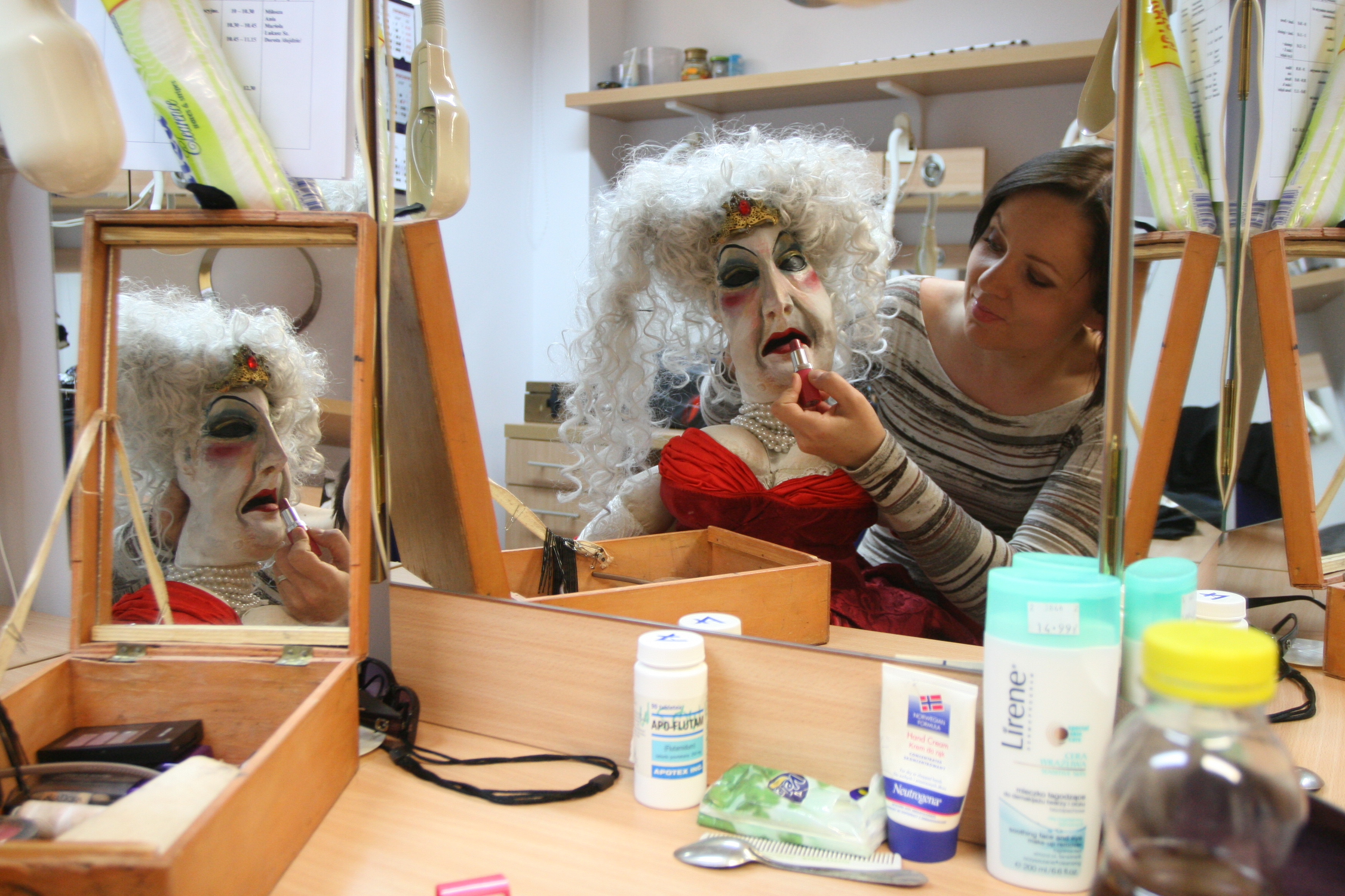 Aktorka siedzi w garderobie, przy biurku z lustrem. Na kolanach trzyma lalkę teatralną, której maluje usta szminką. Lalka odbija sie w lustrze.