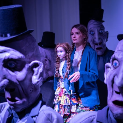 Aktorka stoi pośrodku z lalką Momo. Skupiona, patrzy w jeden punkt. Wokół nich postaci w szarych garniturach i maskach, otaczają Momo.