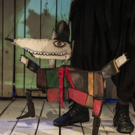 Marionetka wilka na podłodze. Widać sznurki lalki i buty aktora, który ją trzyma.
