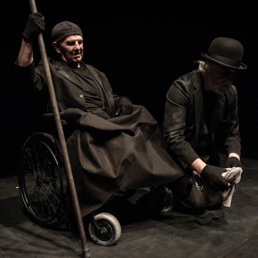 Aktor obwiązuje szmatą goła stopę mężczyzny siedzącego na wózku.
