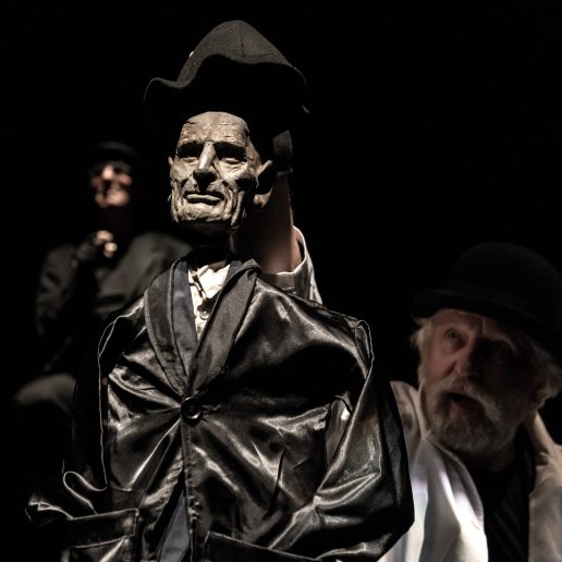 Drewniana, rzeźbiona lalka starszego człowieka w połyskującym czarnym płaszczu. Aktor zakłada mu kapelusz na głowę.