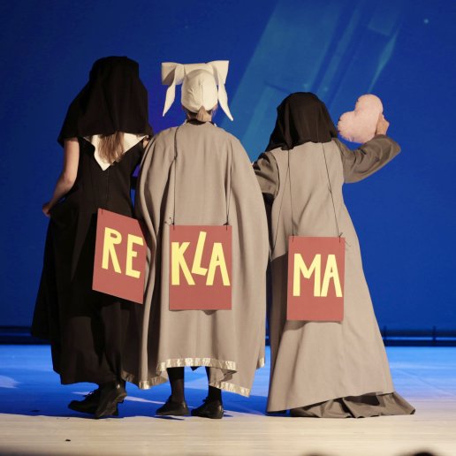 Aktorki w kostiumach z tabliczką z napisem REKLAMA.