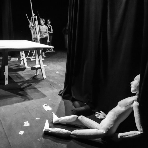 Manekin na scenicznej podłodze oparty o kurtynę. W tle reżyser i aktorzy przy stole i sztalugach.
