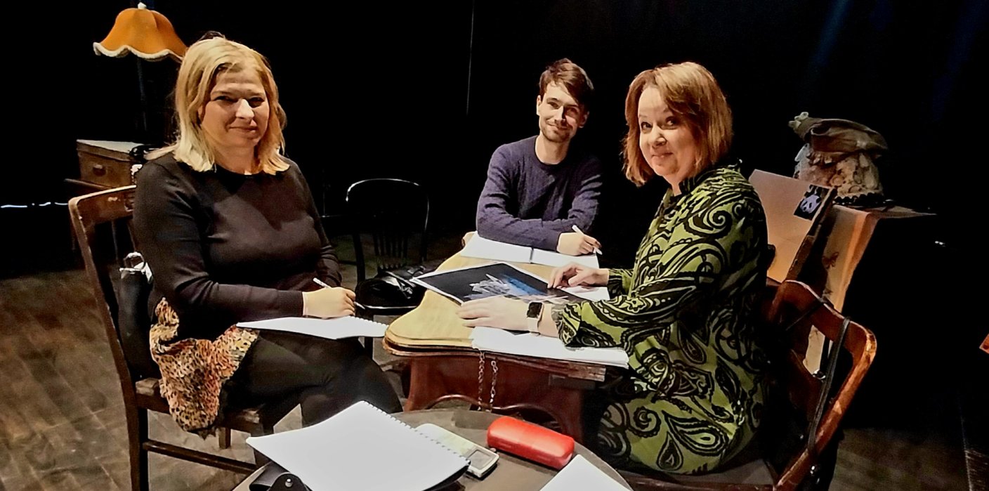 Trzy osoby siedzą przy stole: dwie kobiety, pośrodku mężczyzna. Wszyscy uśmiechnięci. Przed nimi na stole scenariusze sztuki i projekty scenograficzne.