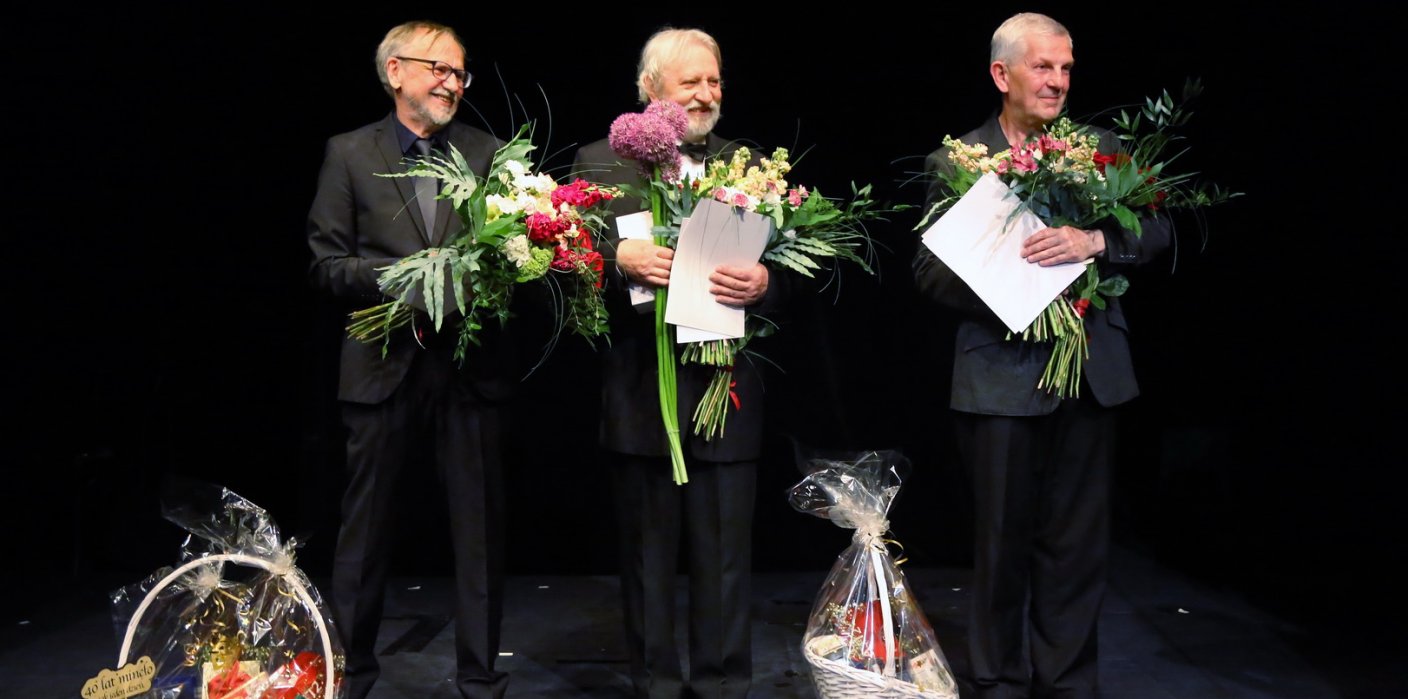 Wszyscy jubilaci: Krystian Kobyłka, Andrzej Mikosza i Andrzej Szymański stoją na scenie, trzymają kwiaty i listy gratulacyjne.